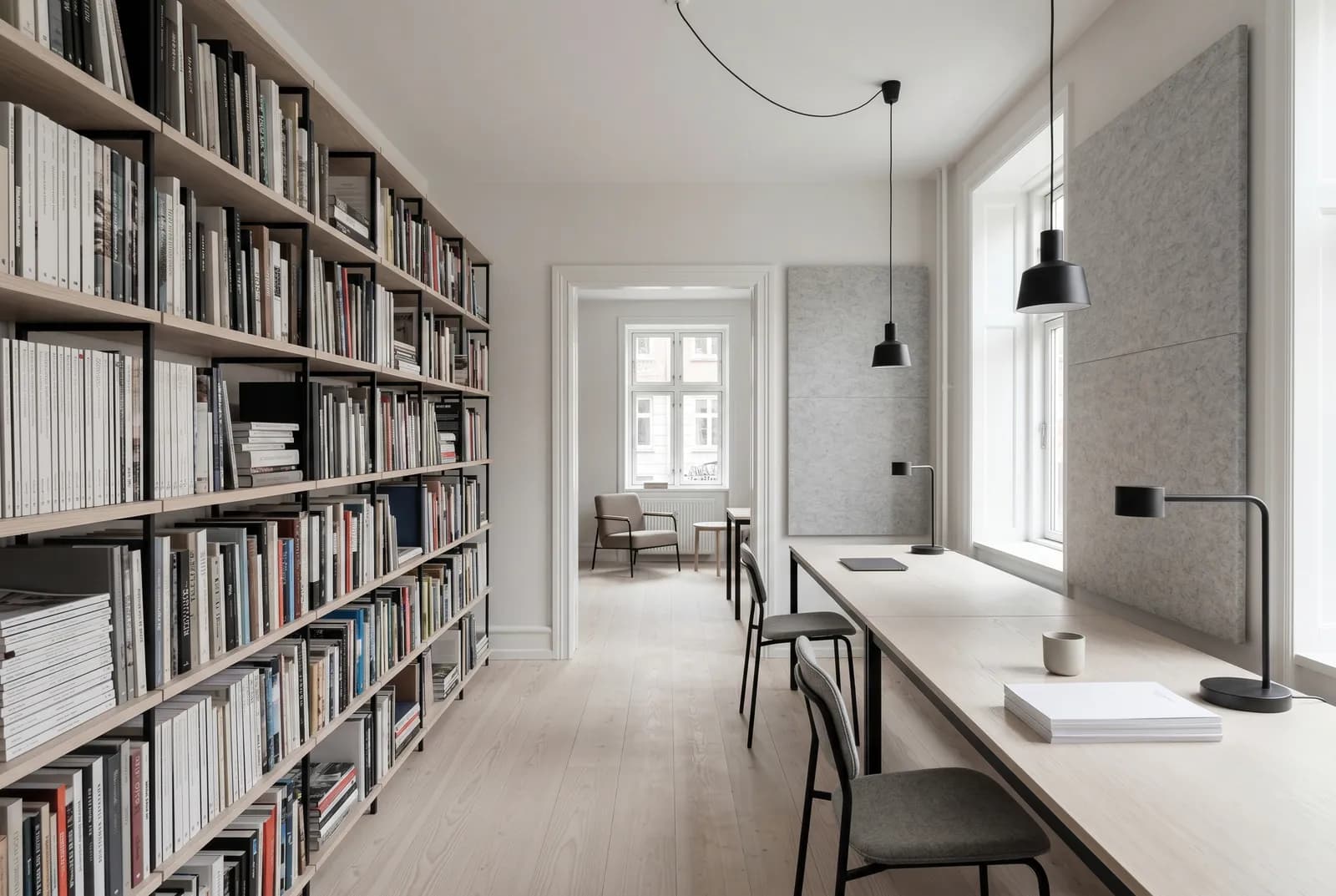 A Copenhagen studio floor with a full-height shared library wall, pale ash desks, and soft Nordic daylight.